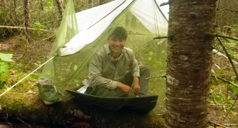 A person smiles from under a tarp shelter covered in a mosquito net